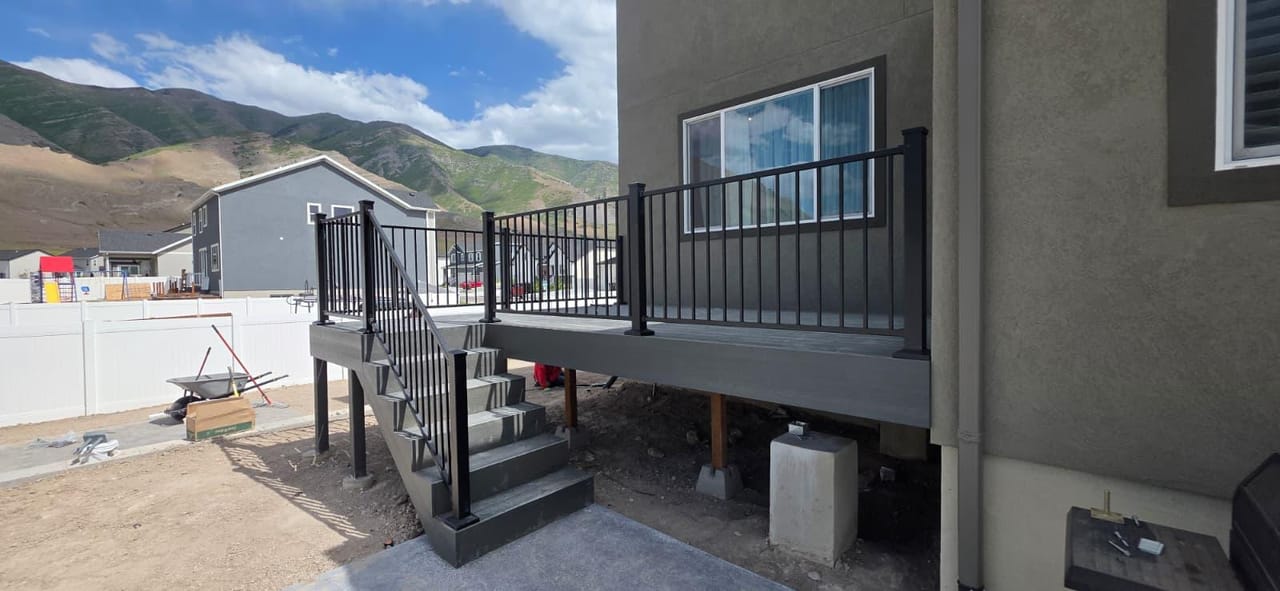 Deck nuevo de composite gris con barandal de metal negro en construcción, escaleras a patio de concreto, con montañas de Utah al fondo — por CasaMás Construction